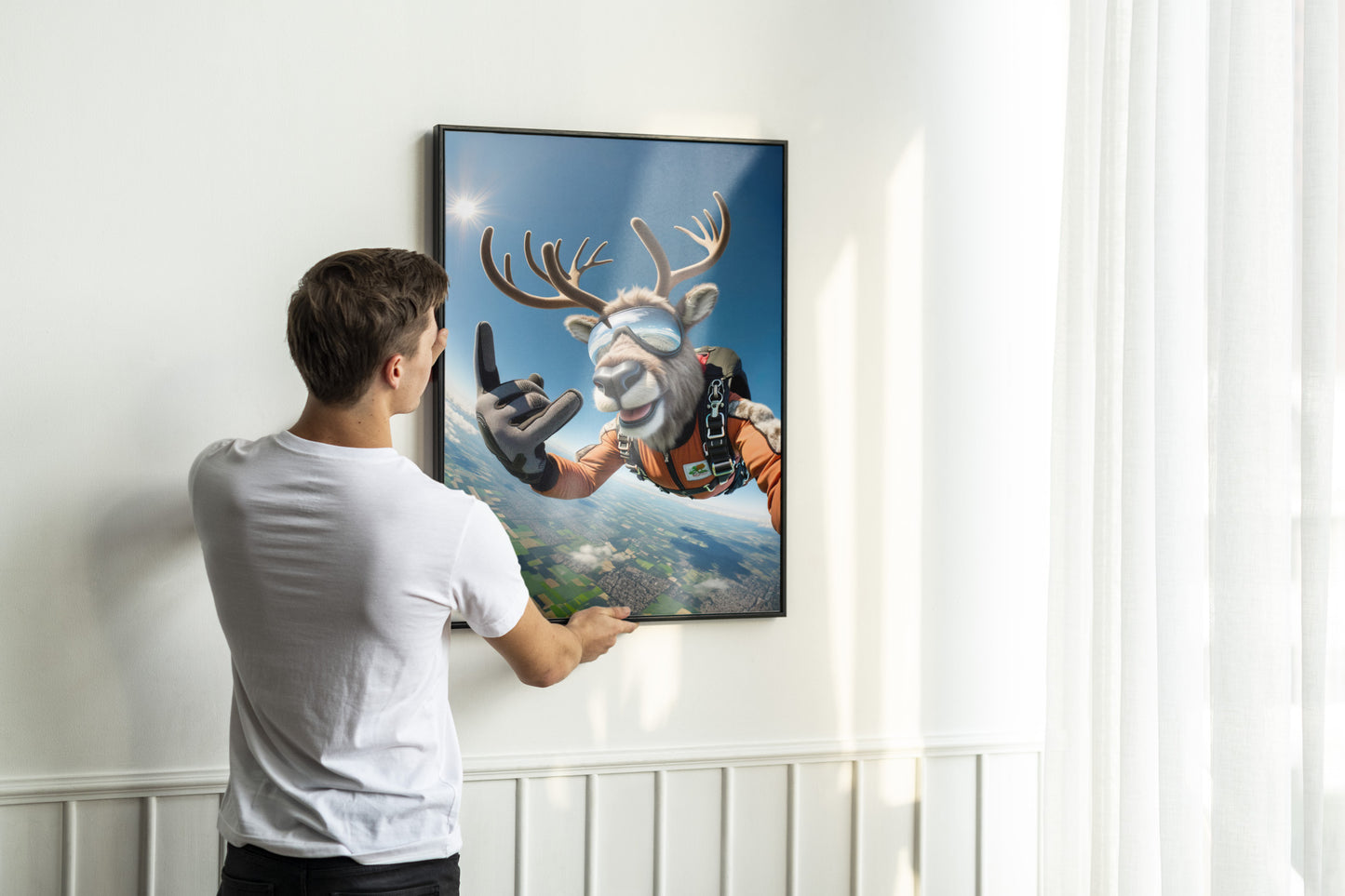 Man in a white T-shirt straightening a black-framed caribou skydiver poster on the wall