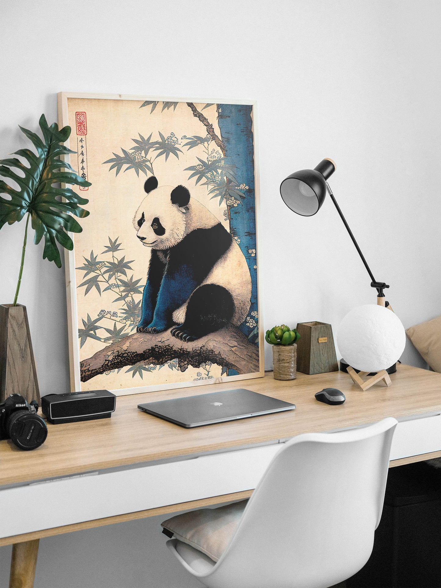 Japanese art panda poster in a natural wood frame leaning on a desk with a black lamp and laptop