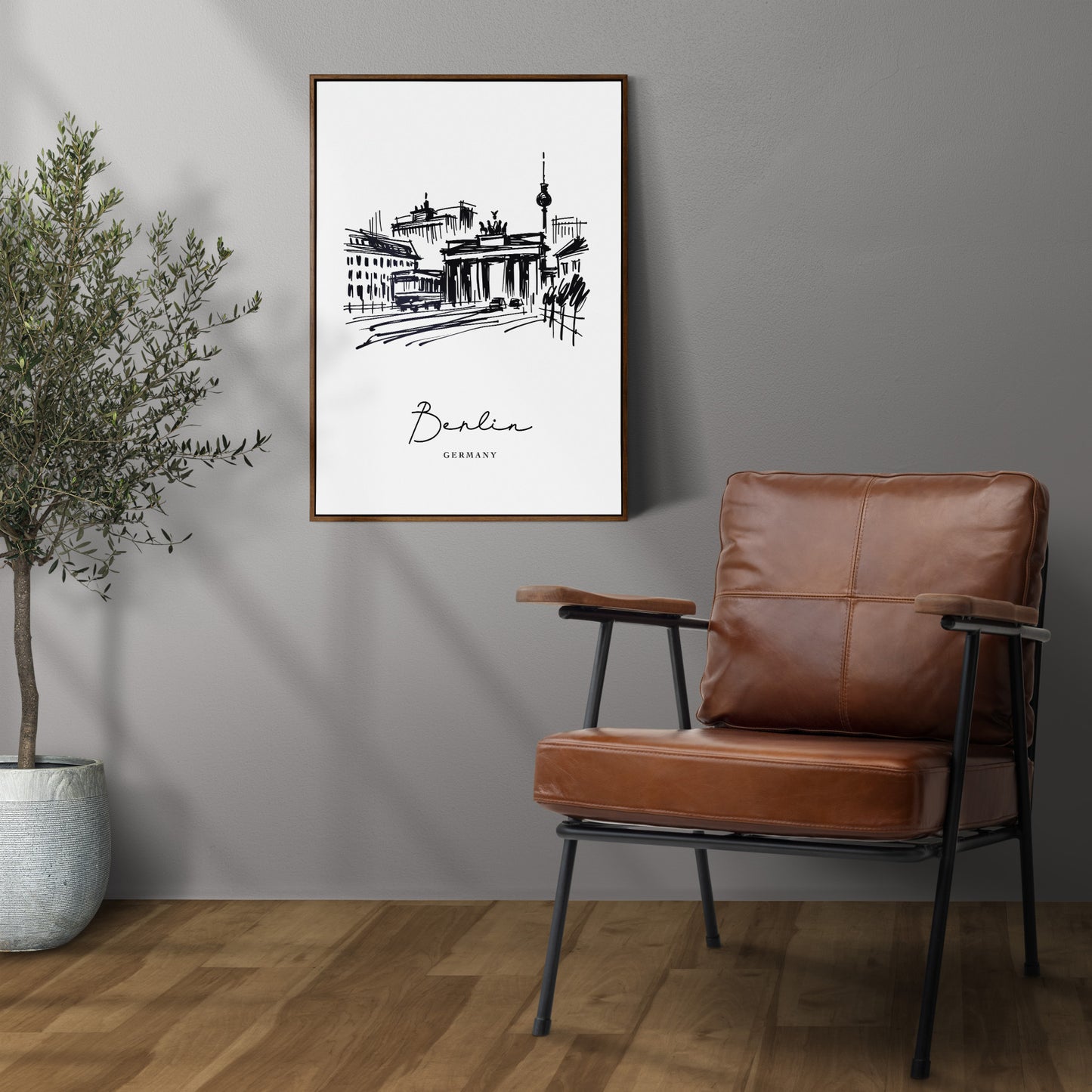 Black and white Berlin poster on a gray wall, dark wood frame above a leather armchair, potted olive tree