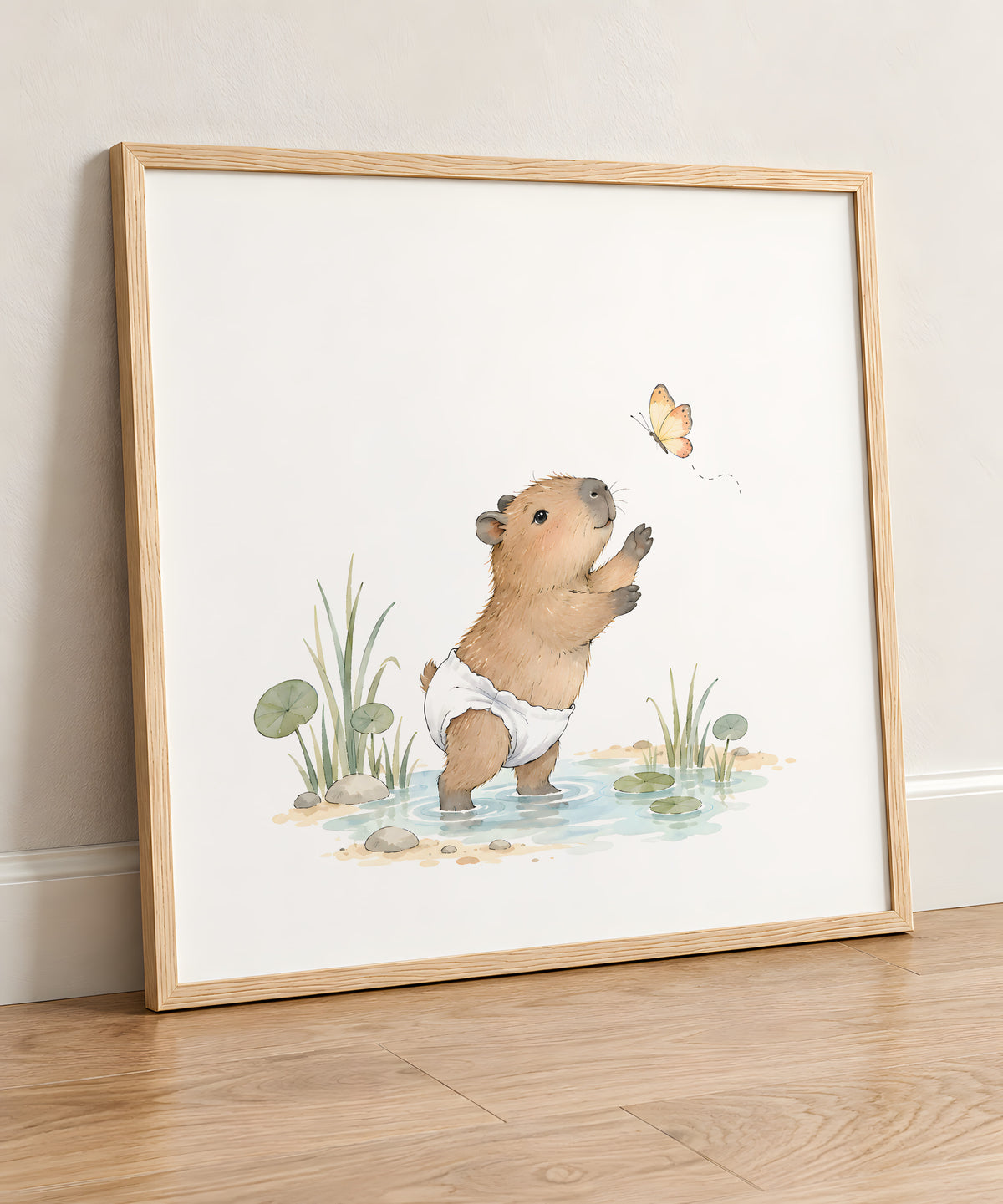 Bébé capybara au bord de l’eau tente d’attraper un papillon : cadre bois clair, 50 × 50 cm, en situation
